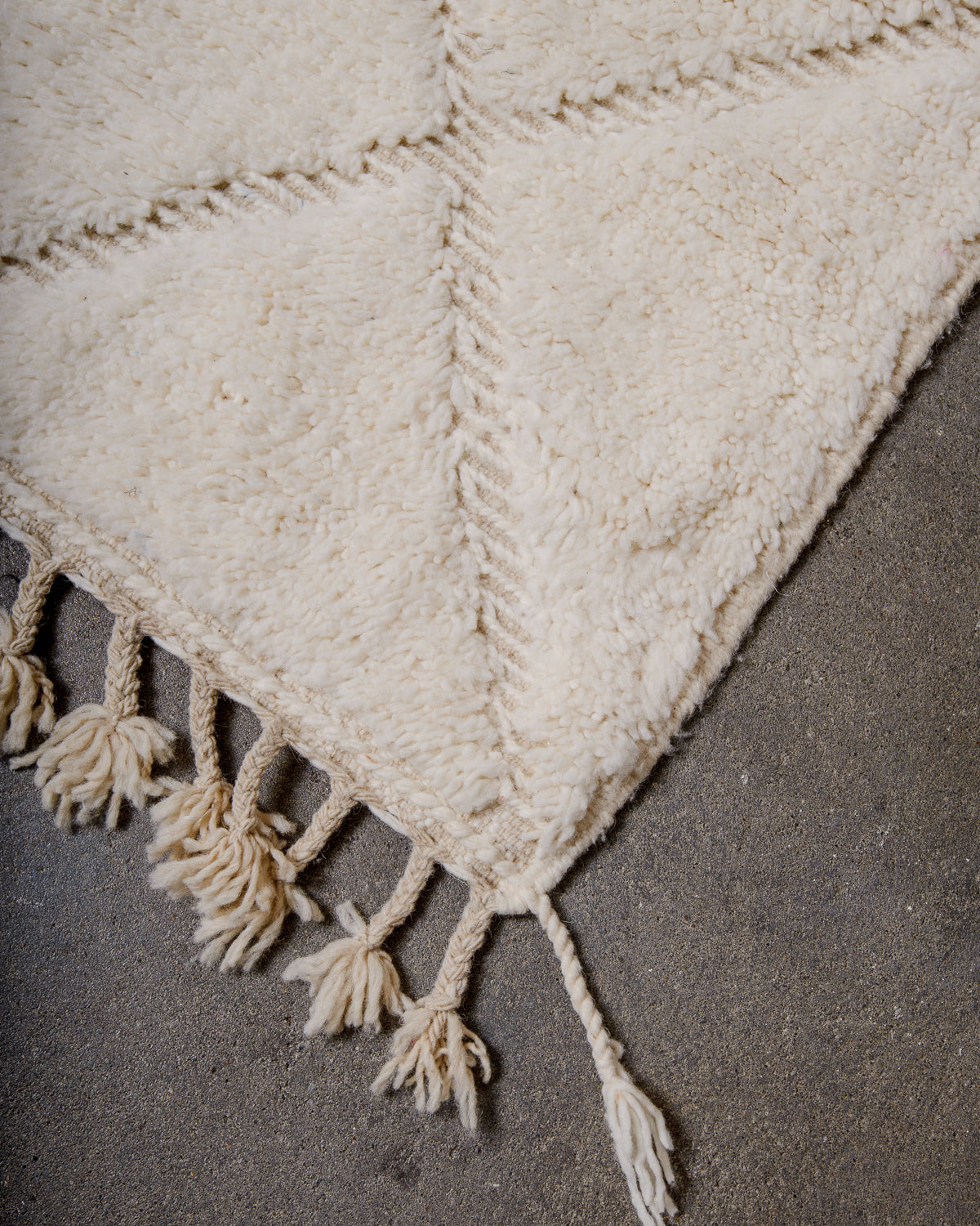 Corner of Berberlin Beniourain rug in cream wool with woven diamond design, laying on concrete floor. 