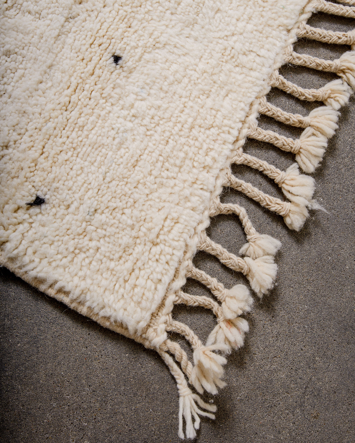Corner of cream Beniourain wool rug with a tightly woven edge and natural wool tassels laying on concrete floor. 