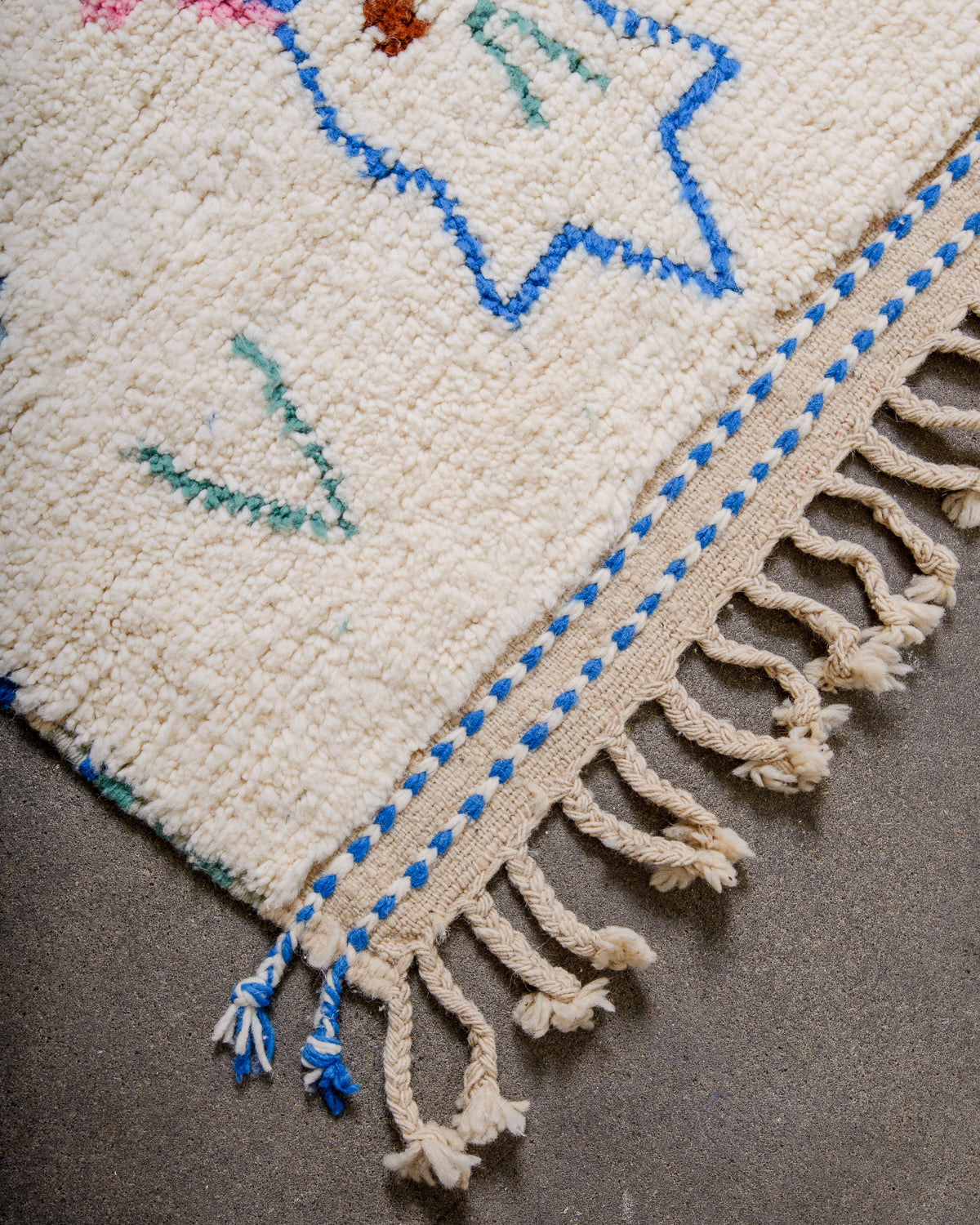 Corner of cream berber rug that is finished with tightly woven ends that also have two blue and cream wool woven lines running across them.