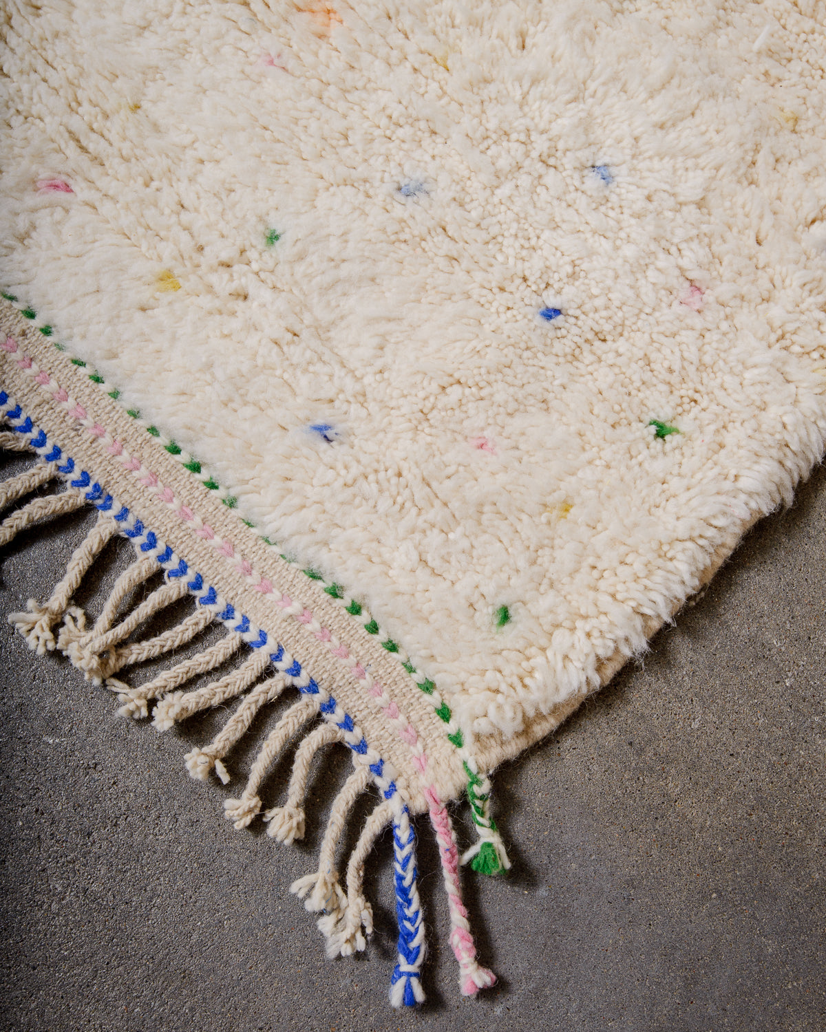 Corner of Berber rug with green, Blue and pink stitching and natural wool tassels across the top