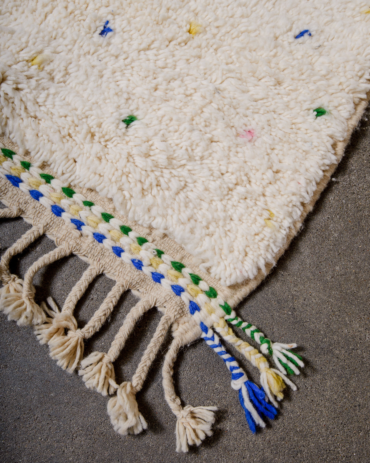 Corner of Berber rug with green, Blue and yellow stitching and natural wool tassels across the top.