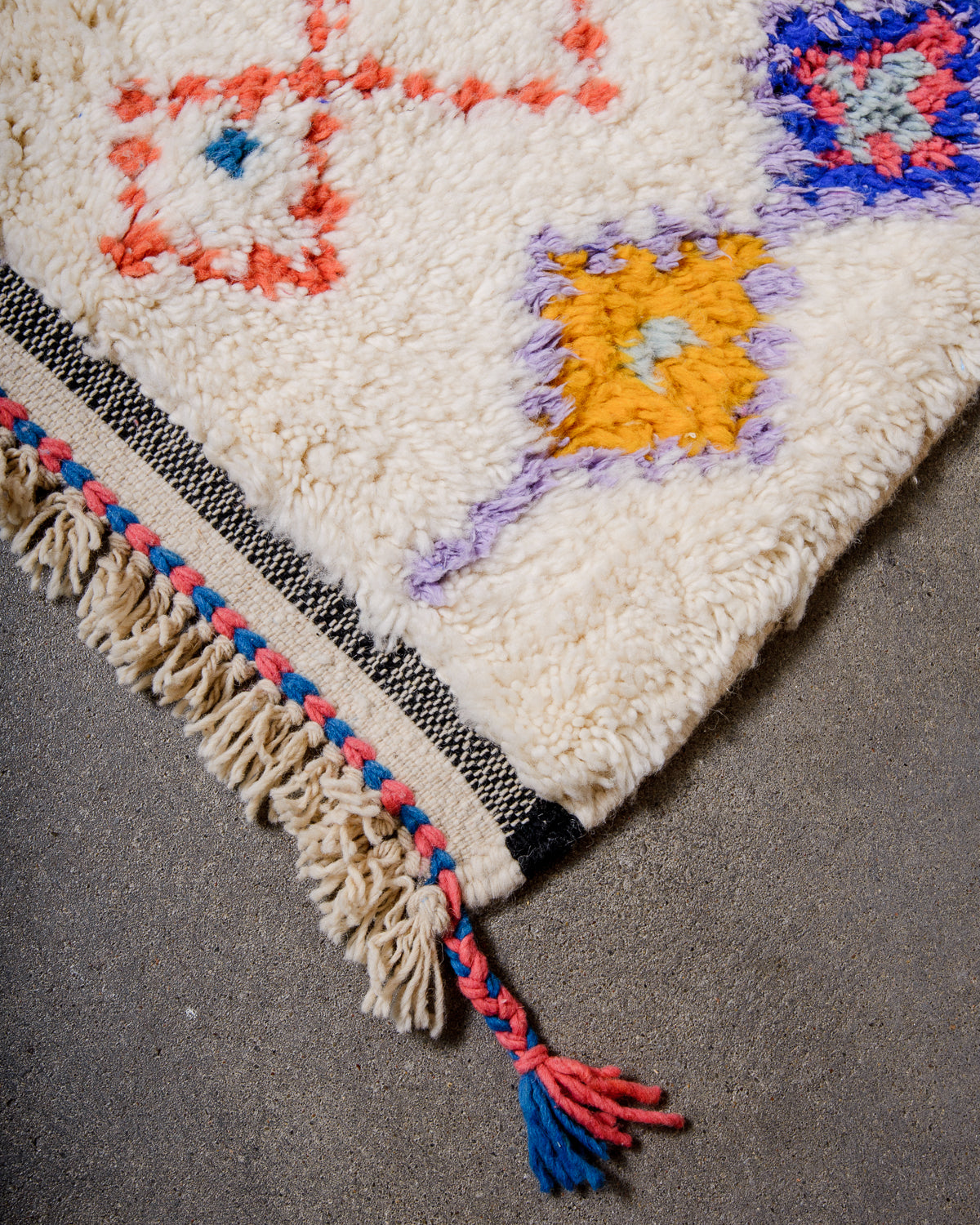 Corner of Beniourain rug with cream wool and design elements in lavender, yellow, coral and blue, with a tightly woven end in black and cream and natural wool tassels.