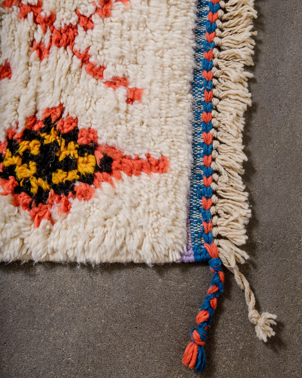Corner of Beniourain rug with cream wool and design elements in lavender, yellow, coral and blue, with a tightly woven end in blue and coral, and natural wool tassels.