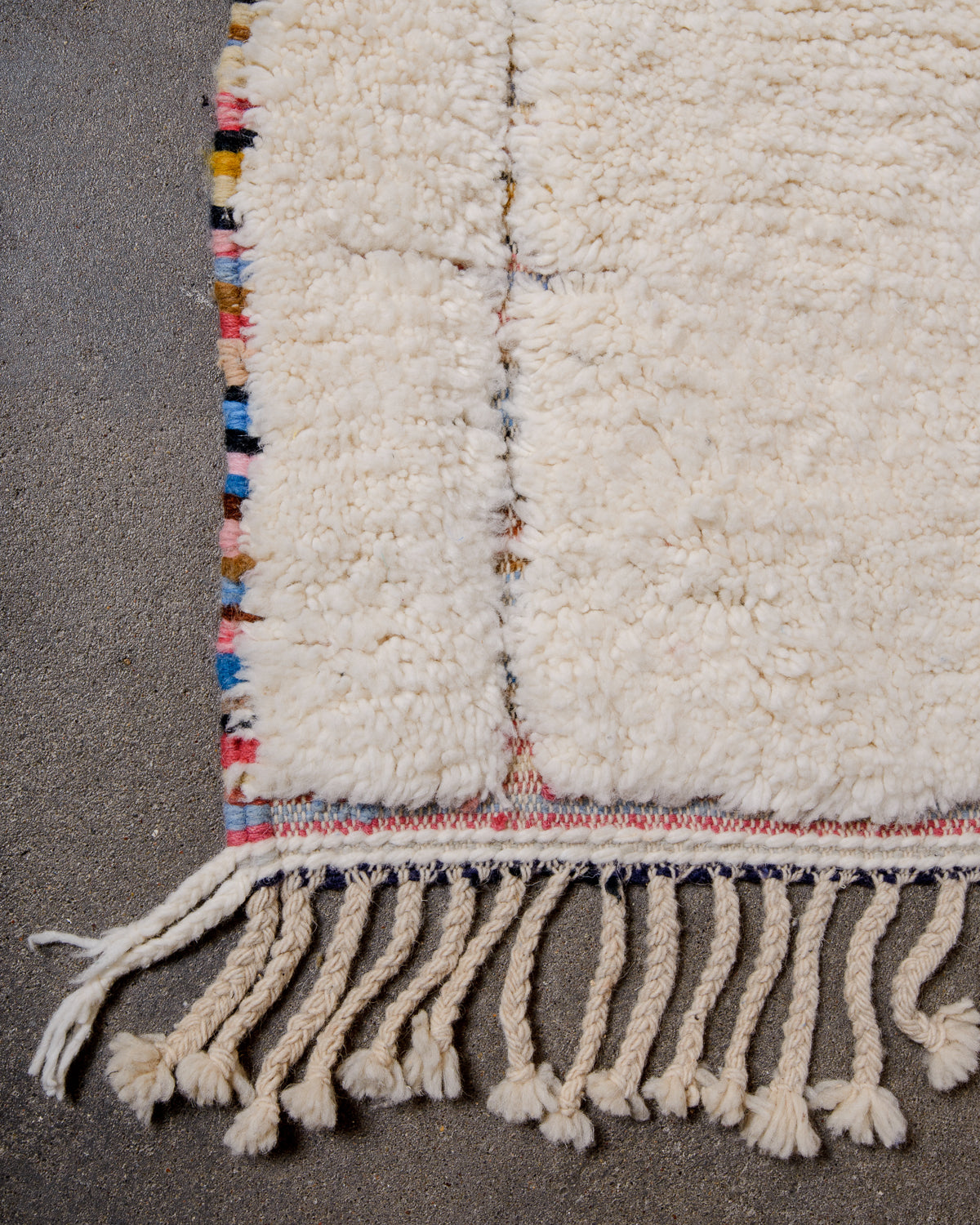 Corner of rug with natural wool tassels, cream wool pile and multicoloured base that shows in the parts of the rug that are not knotted and on the edge. 