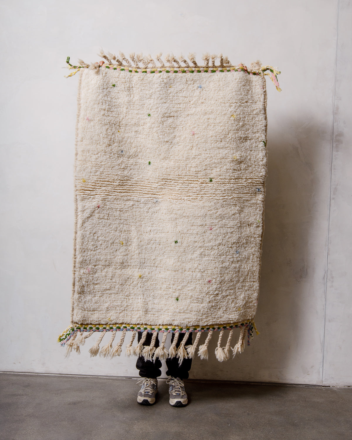 Small Berberlin Beniourain rug in cream with coloured dots and edge, being held up by a person in front of a white wall. 