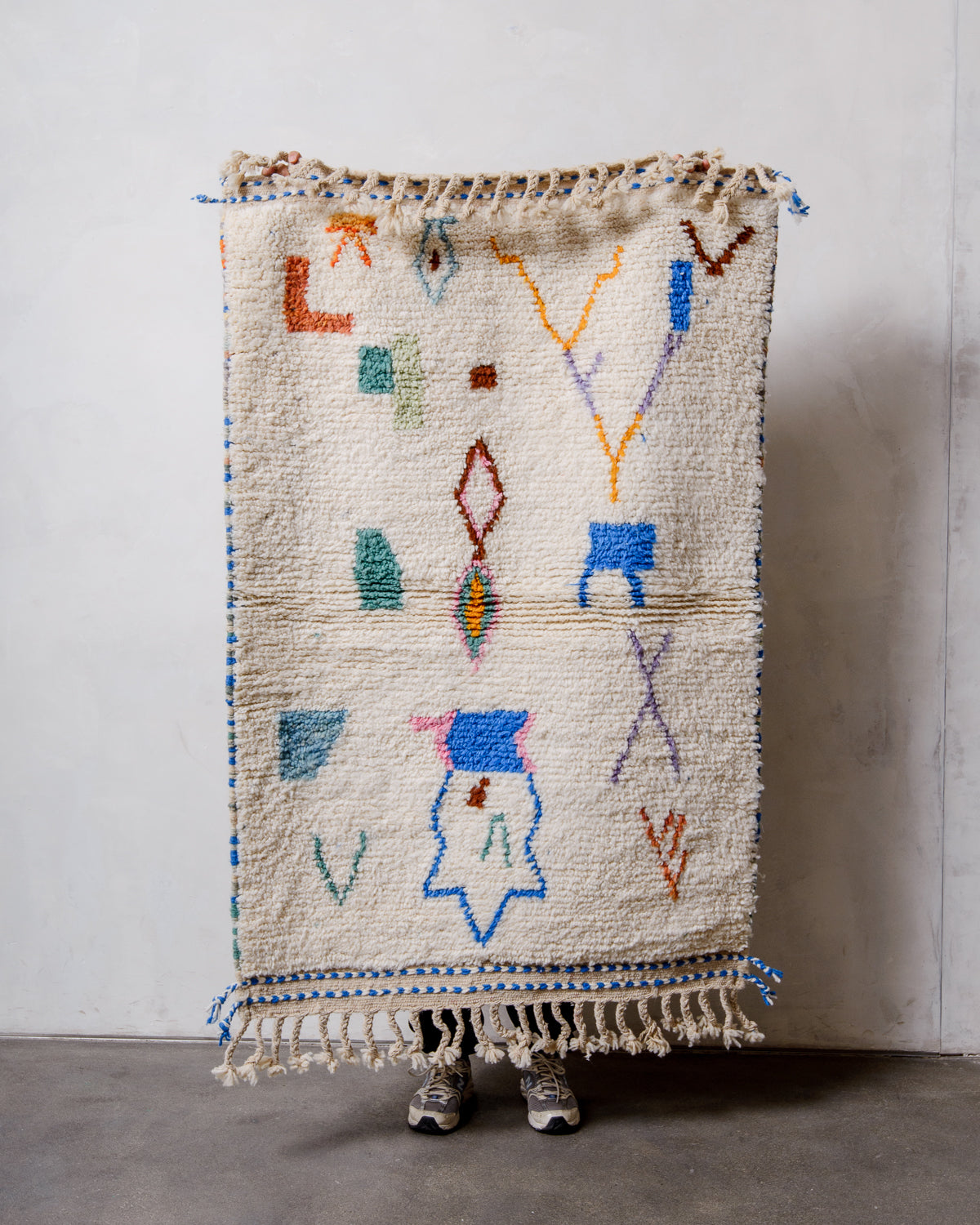 Small Berberlin Beniourain rug made of cream wool with colourful, abstract design, being held up in front of a white wall. 