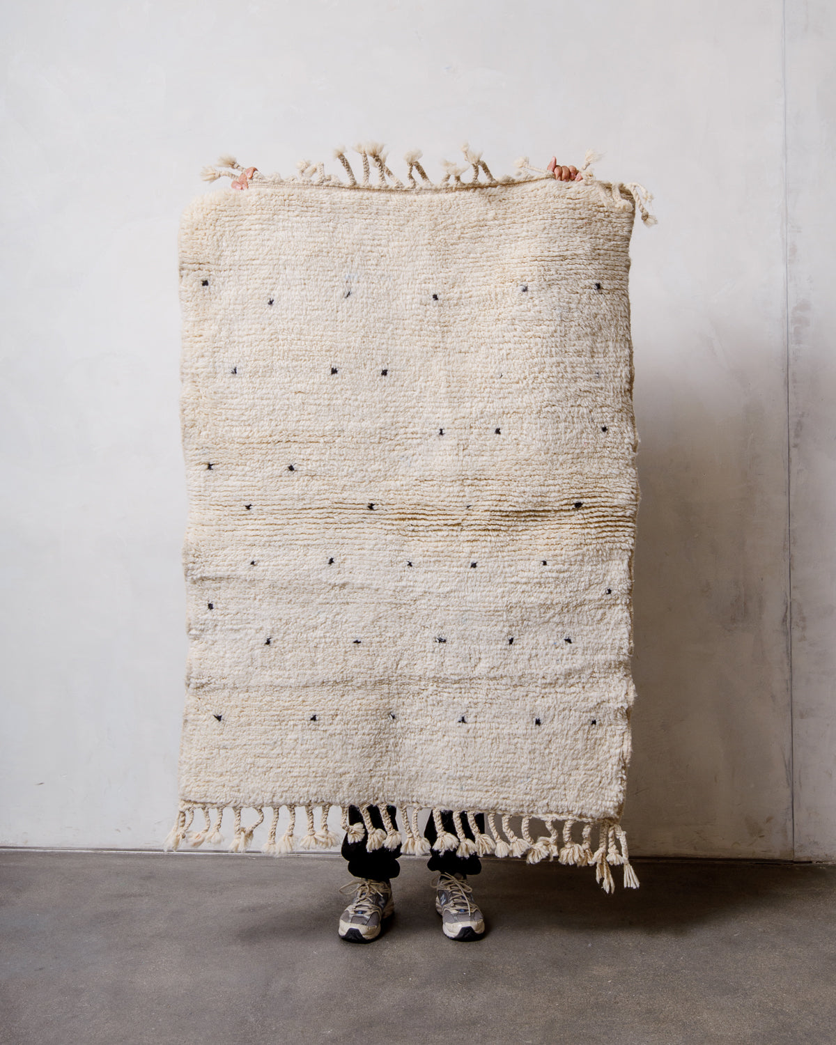 Small Berberlin Beniourain rug in cream wool with black dots, being held up by a person in front of a white wall. 