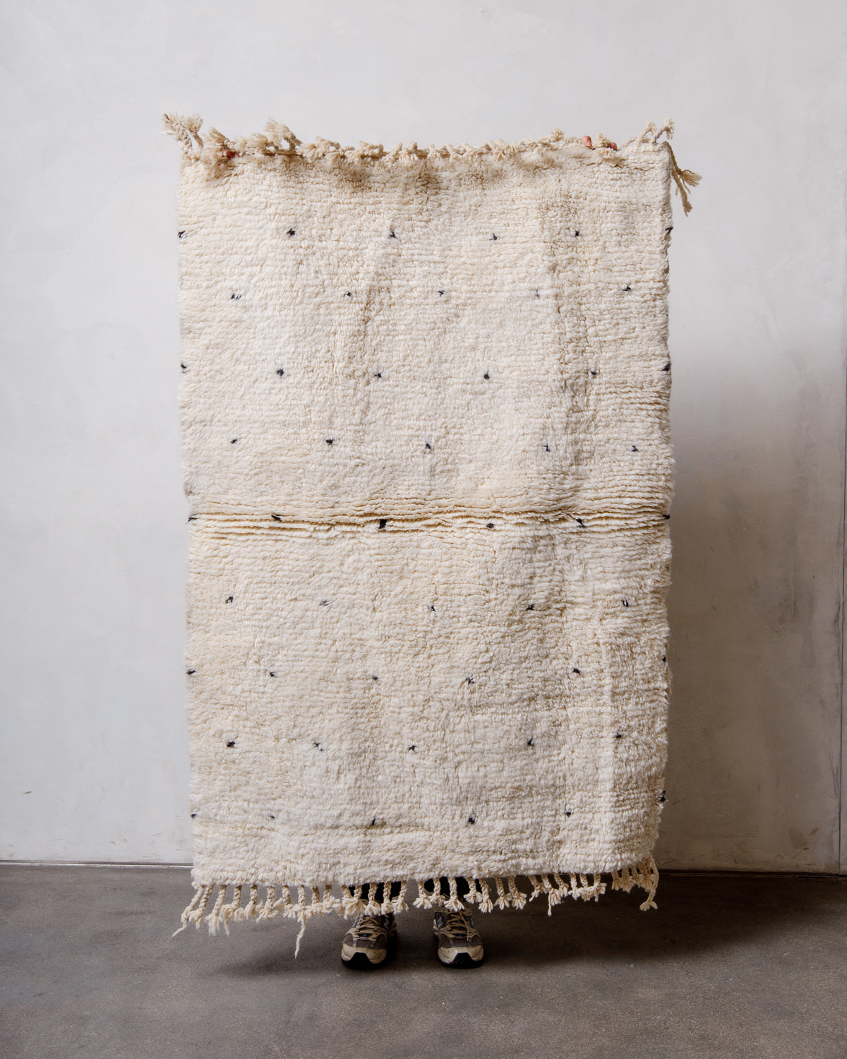 Small Berberlin Beniourain rug in cream wool with black dots, being held up by a person in front of a white wall. 