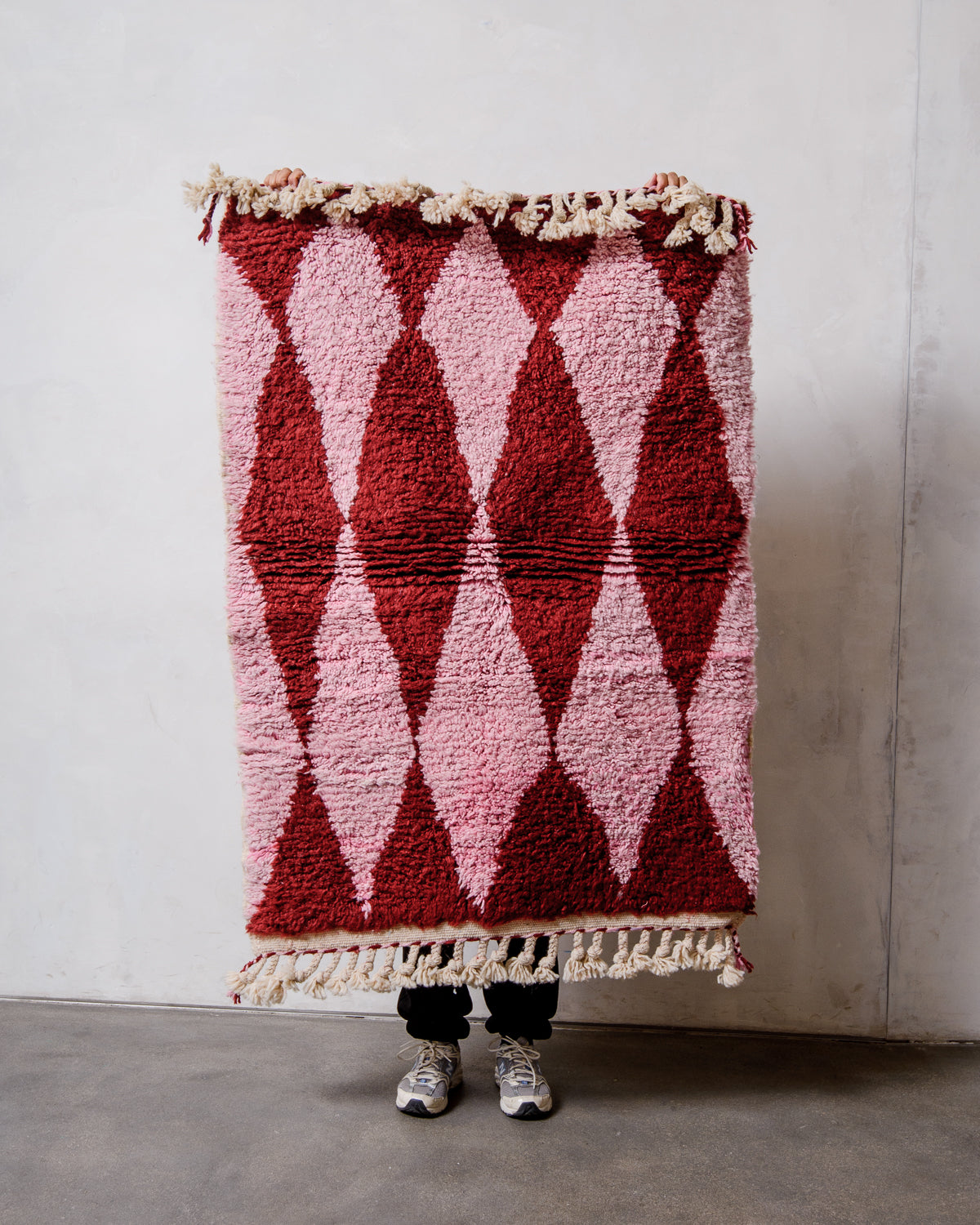 Small Beniourain rug with a harlequin pattern in pink and burgundy wool and with natural wool tassels, being held up in front of a white wall. 