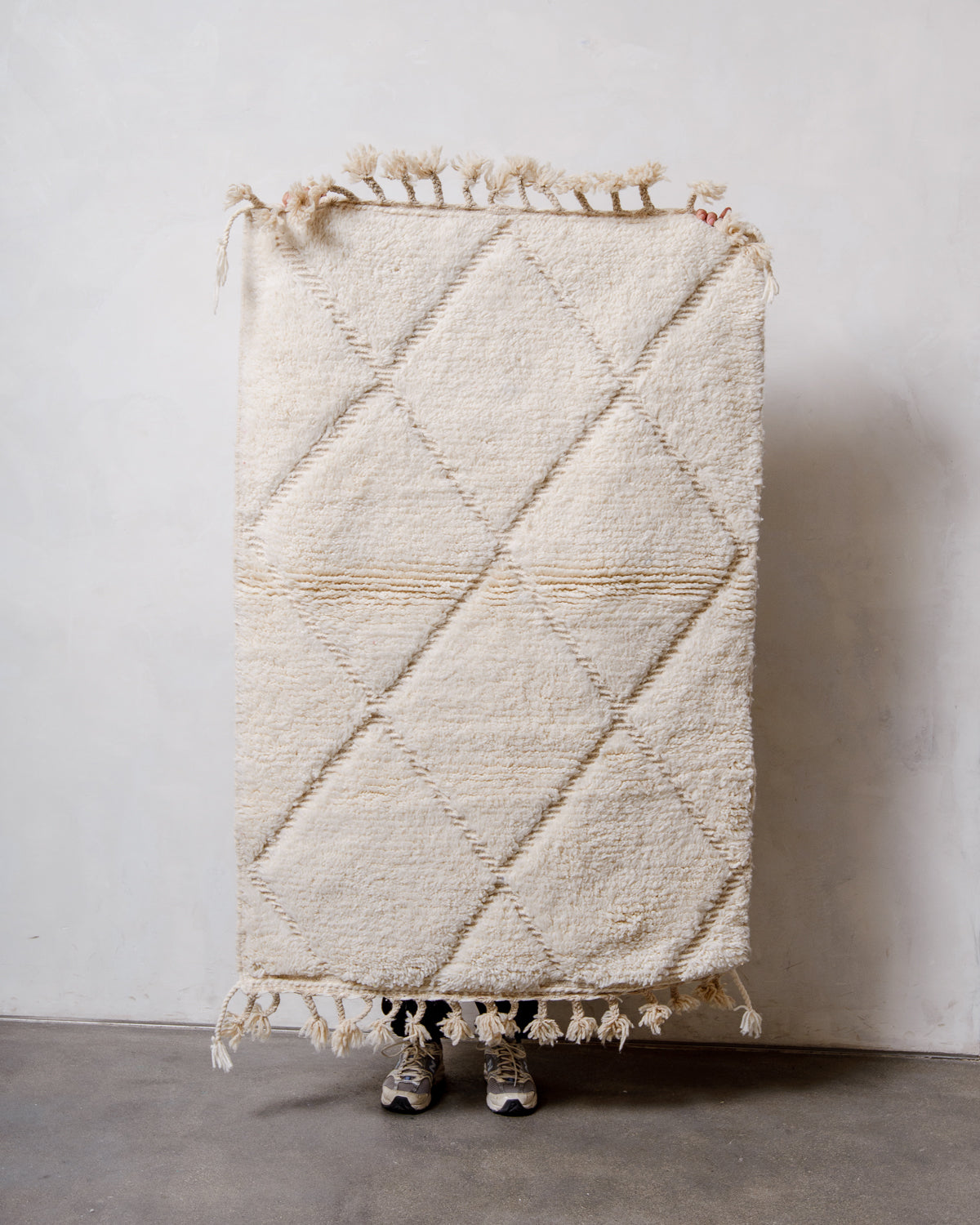 Berberlin Beniourain rug in cream wool with woven diamond design, being held up in front of a white wall. 