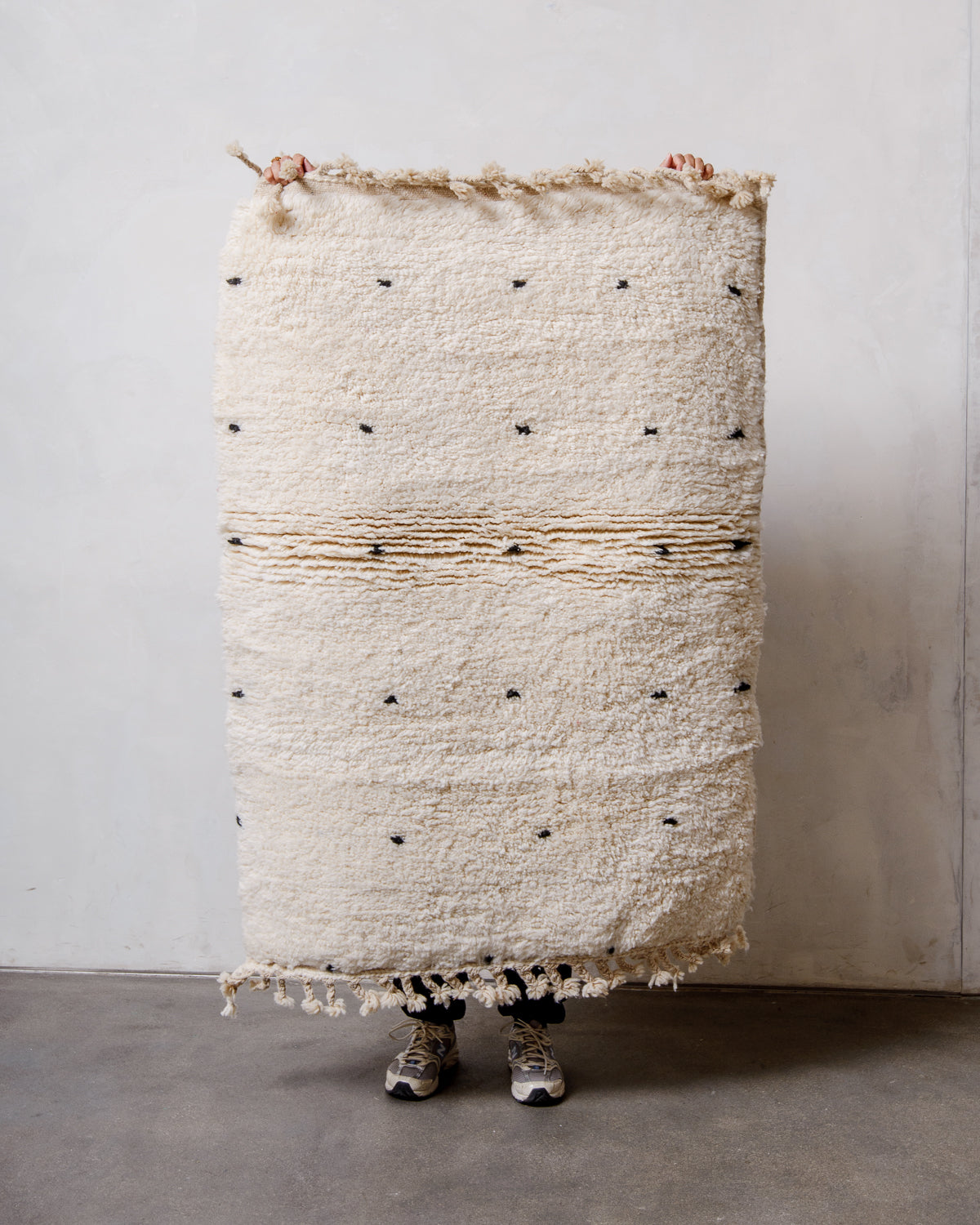Small Berberlin Beniourain rug in cream wool with large black dots, being held up by a person in front of a white wall. 