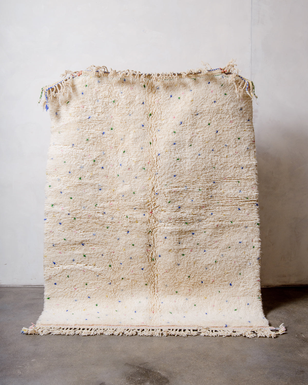 Berberlin Beniourain rug in cream wool with colourful dots being held up in front of a white wall. 
