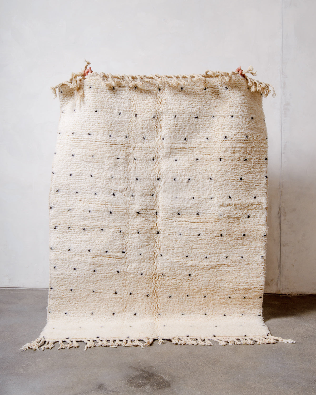Berberlin Beniourain rug in cream wool with black dots being held up in front of a white wall. 
