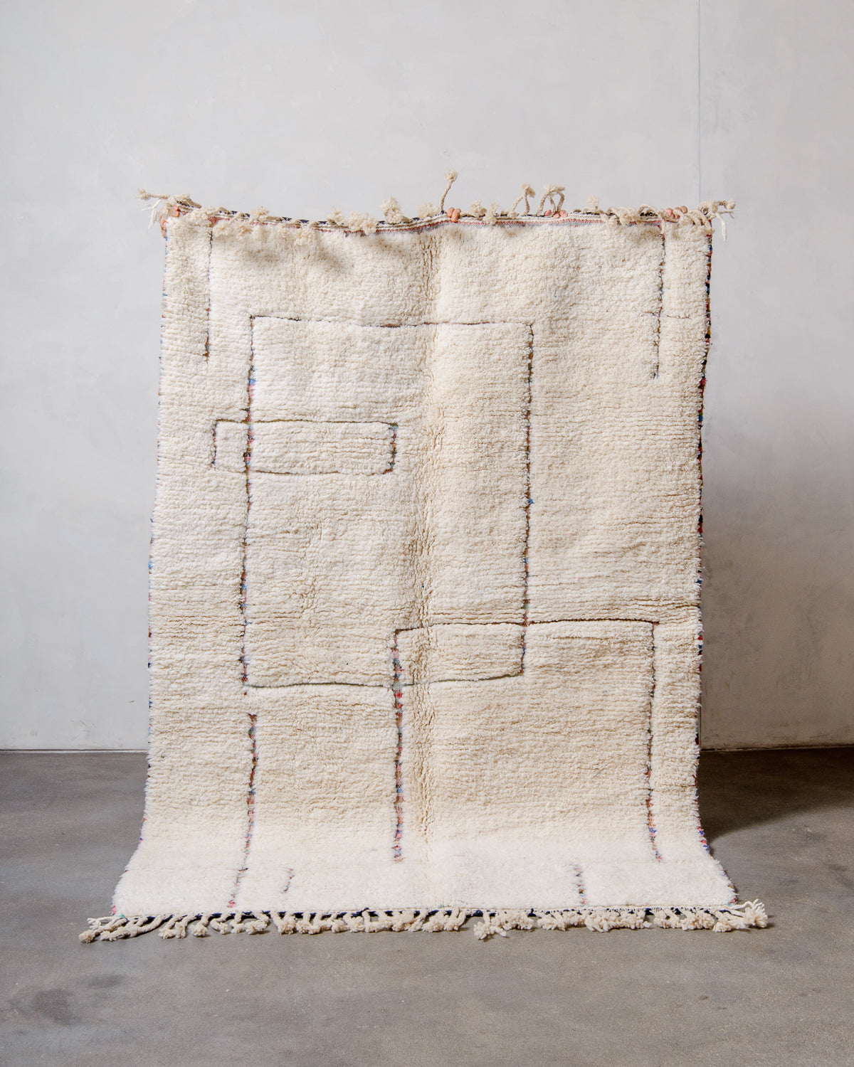 Cream wool rug with a geometric design in multi-coloured wool being held up in front of a wall. 