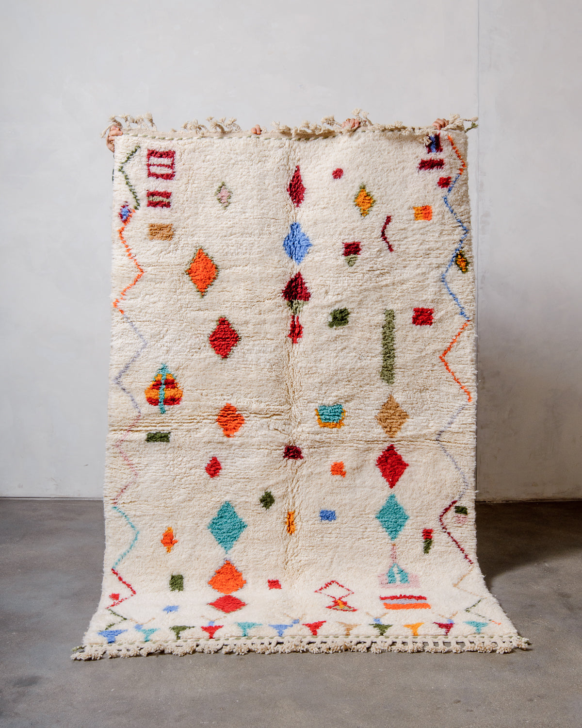Berberlin Beniourain rug in colourful wool and cream, being held up in front of a white wall. 