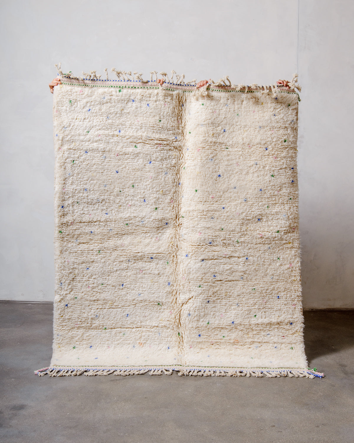 Berberlin Beniourain rug in cream wool with coloured dots being held up in front of a white wall. 