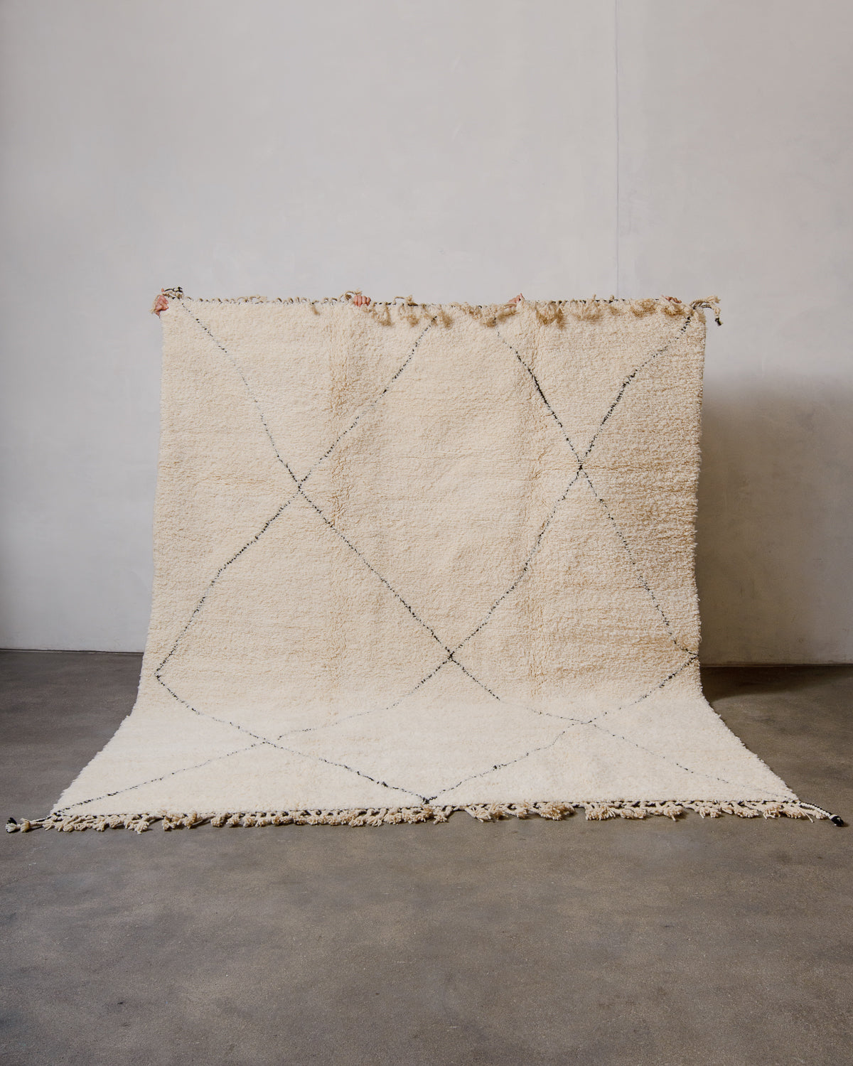 Berberlin Beniourain rug in cream wool with tassels across the top and bottom edges, being held up against a white wall. Large size rug. 