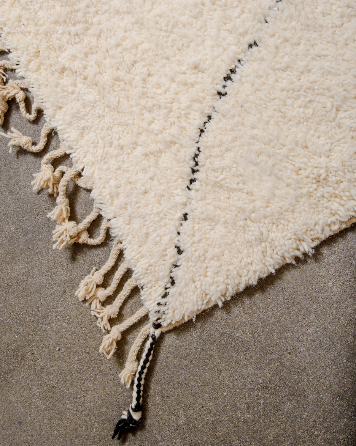 Corner of berber rug showing black and white corner tassel and natural wool tassels across the top edge.