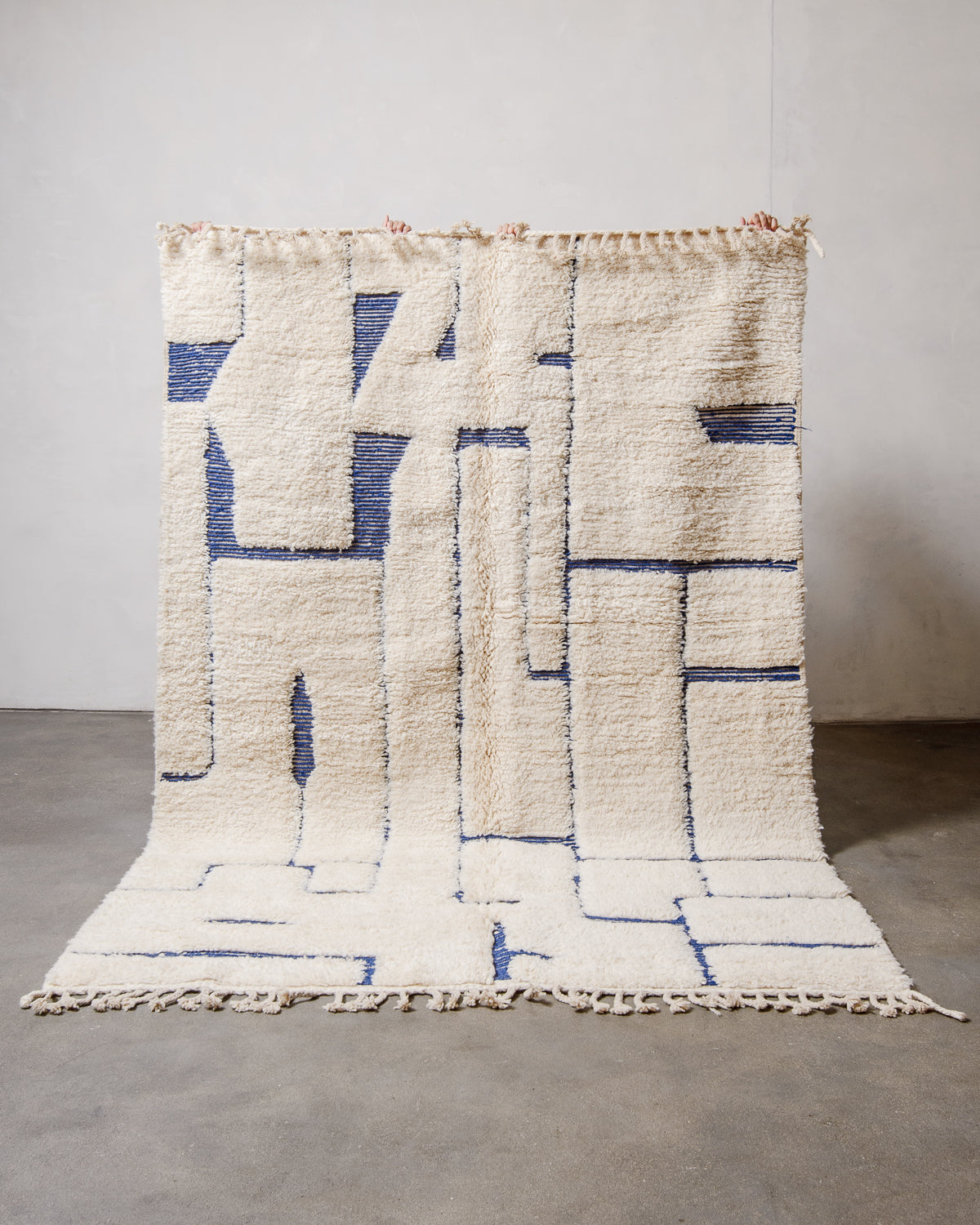 Berberlin Beniourain Rug made of cream and blue wool, held up in front of a white wall. 