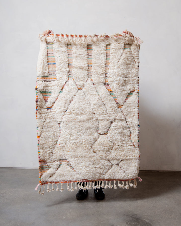Small Berberlin Beniourain rug with cream wool pile and woven base of multicoloured stripes, being held up in front of a white wall. 