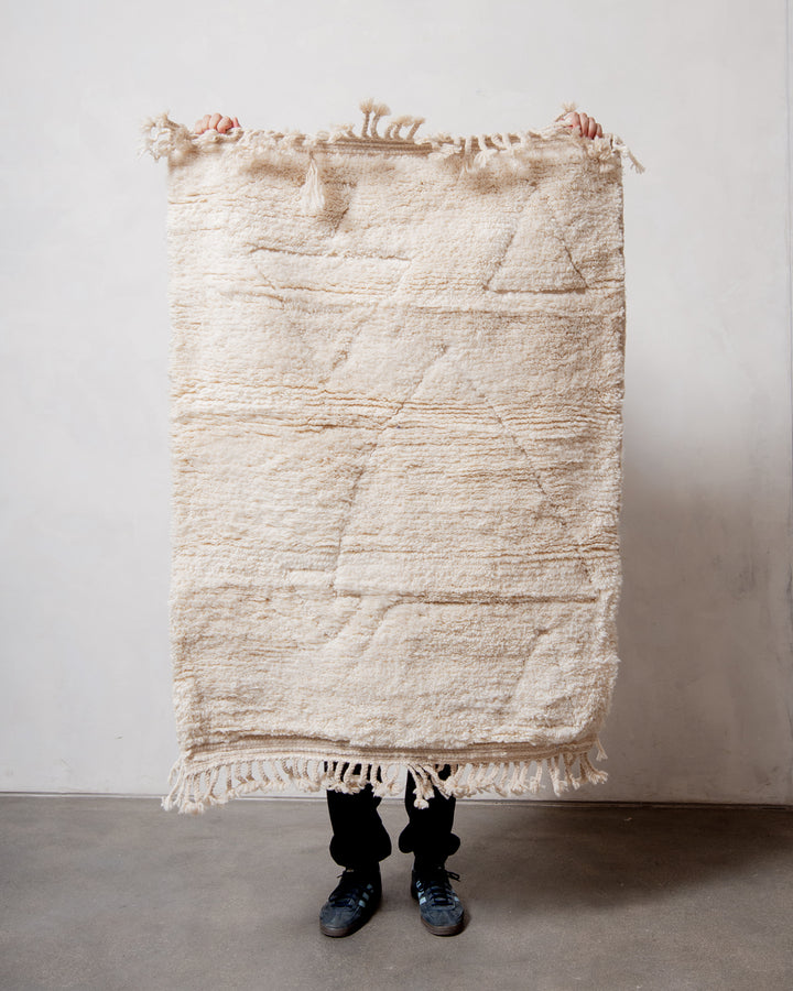 Small Berberlin Beniourain rug in cream wool with design of triangles, being held up in front of a white wall. 