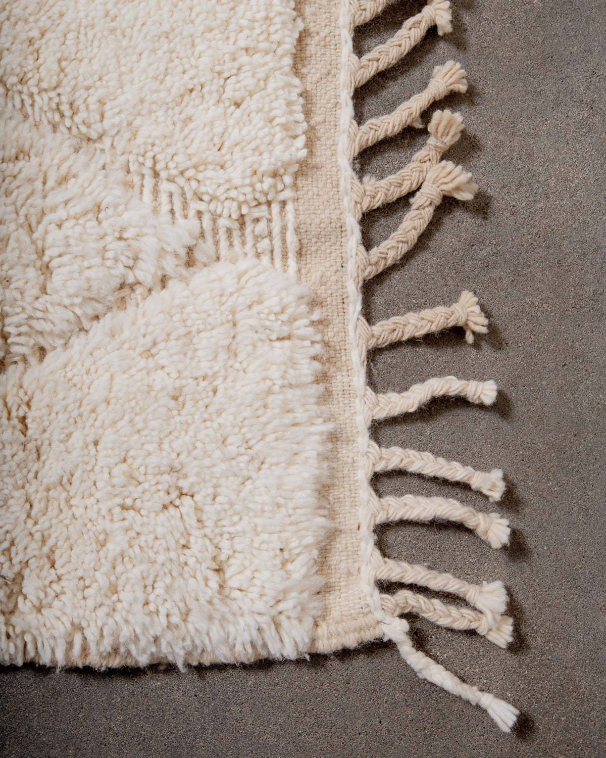Corner of cream Beniourain wool rug with a tightly woven edge and natural wool tassels laying on concrete floor. 