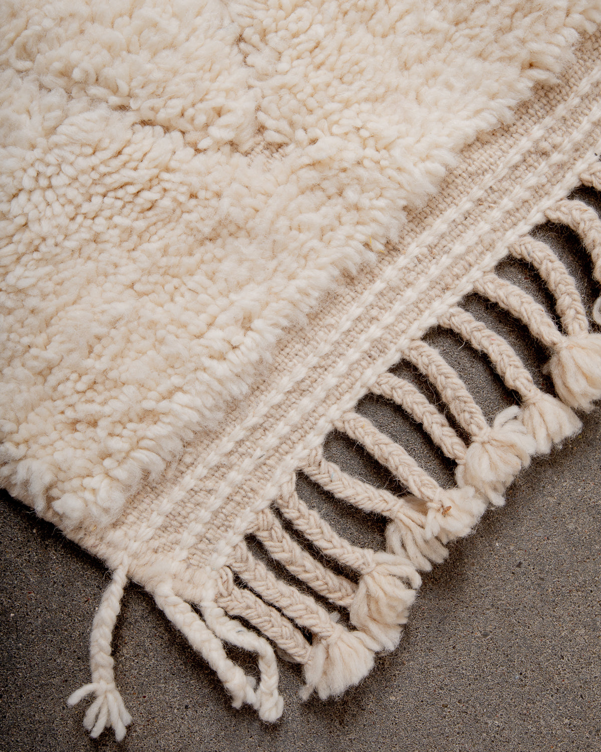 Corner of cream Beniourain wool rug with a tightly woven edge and natural wool tassels laying on concrete floor. 