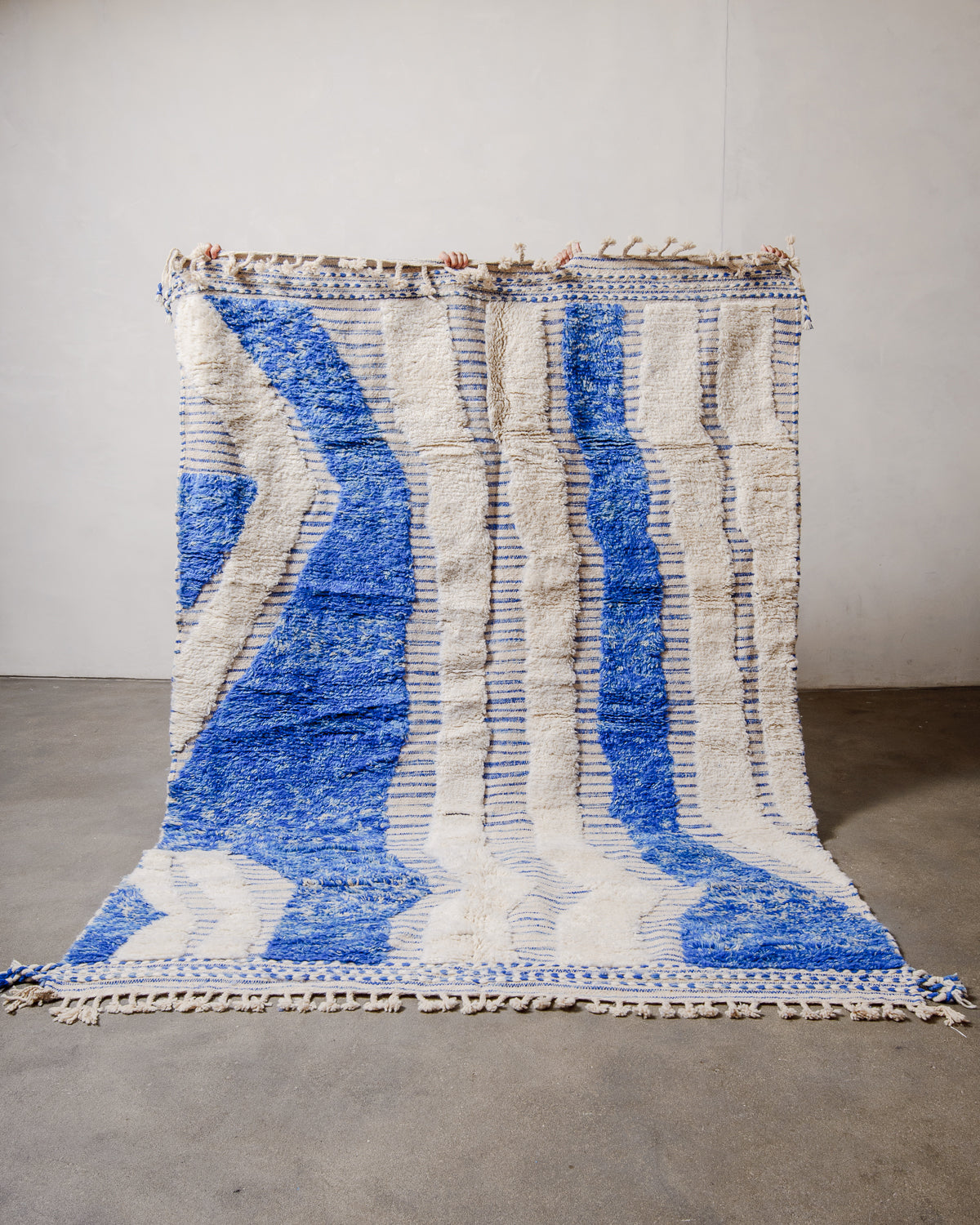 Berberlin Beniourain Rug made of cream and blue wool, held up in front of a white wall. 