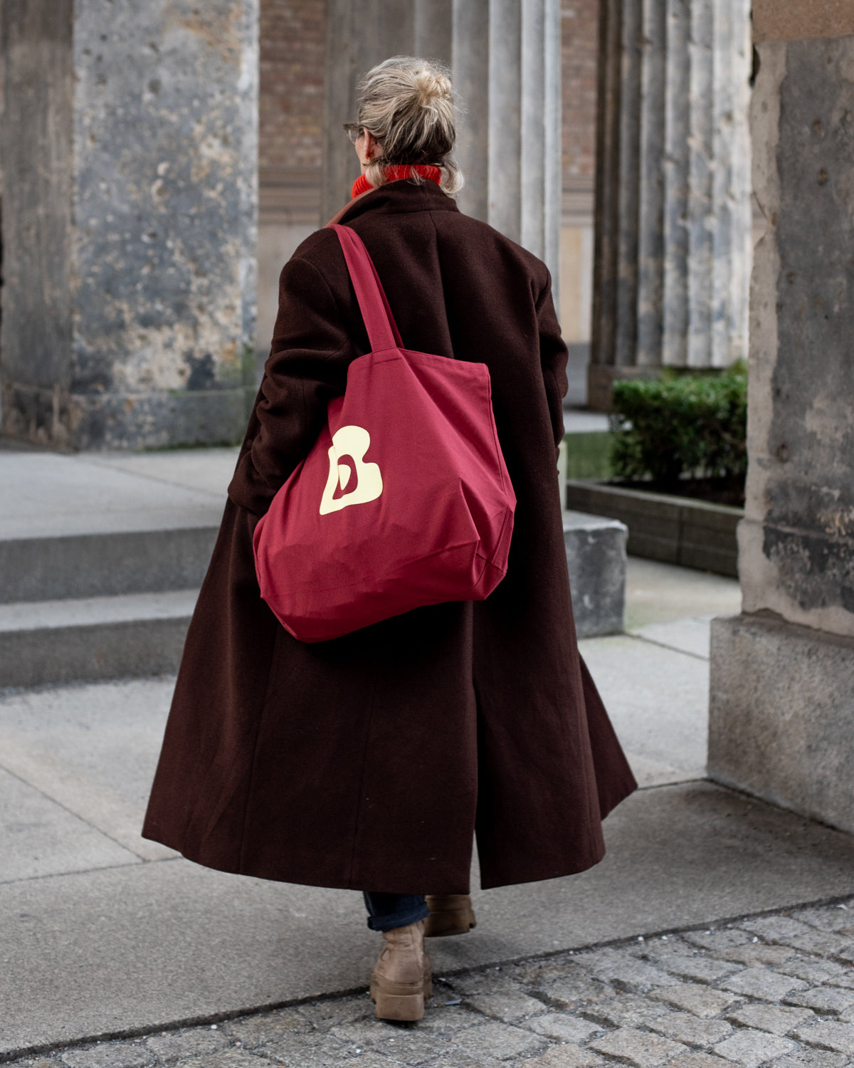 Woman carrying burgundy tote bag with lemon Berberlin logo over shoulder and walking between museum columns.