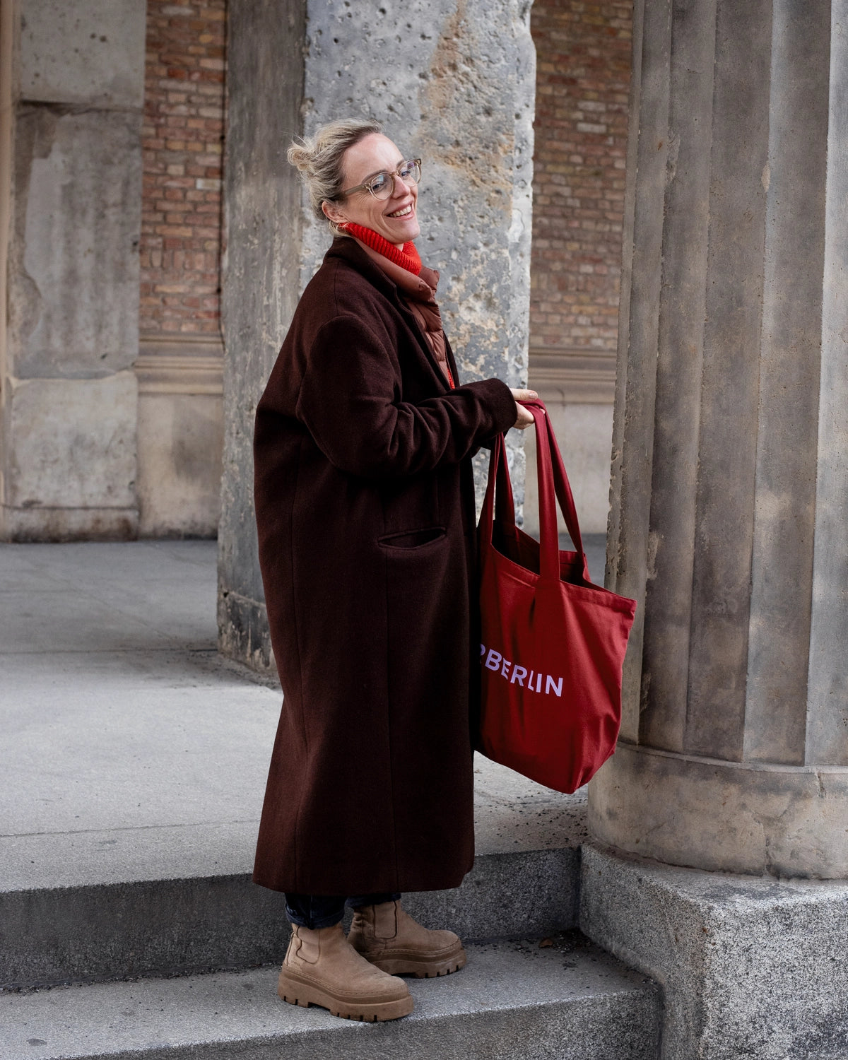 Woman in long brown coat smiling and holding burgundy canvas tote bag with lilac writing on it.