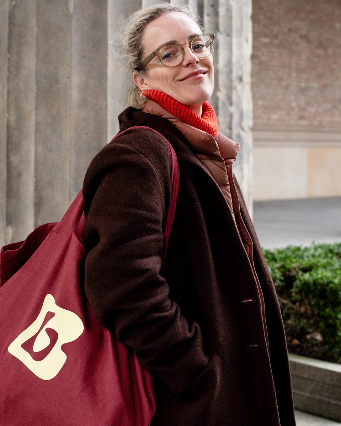Woman carrying burgundy tote bag with lemon Berberlin logo and wearing a brown wool coat standing in front of museum columns.
