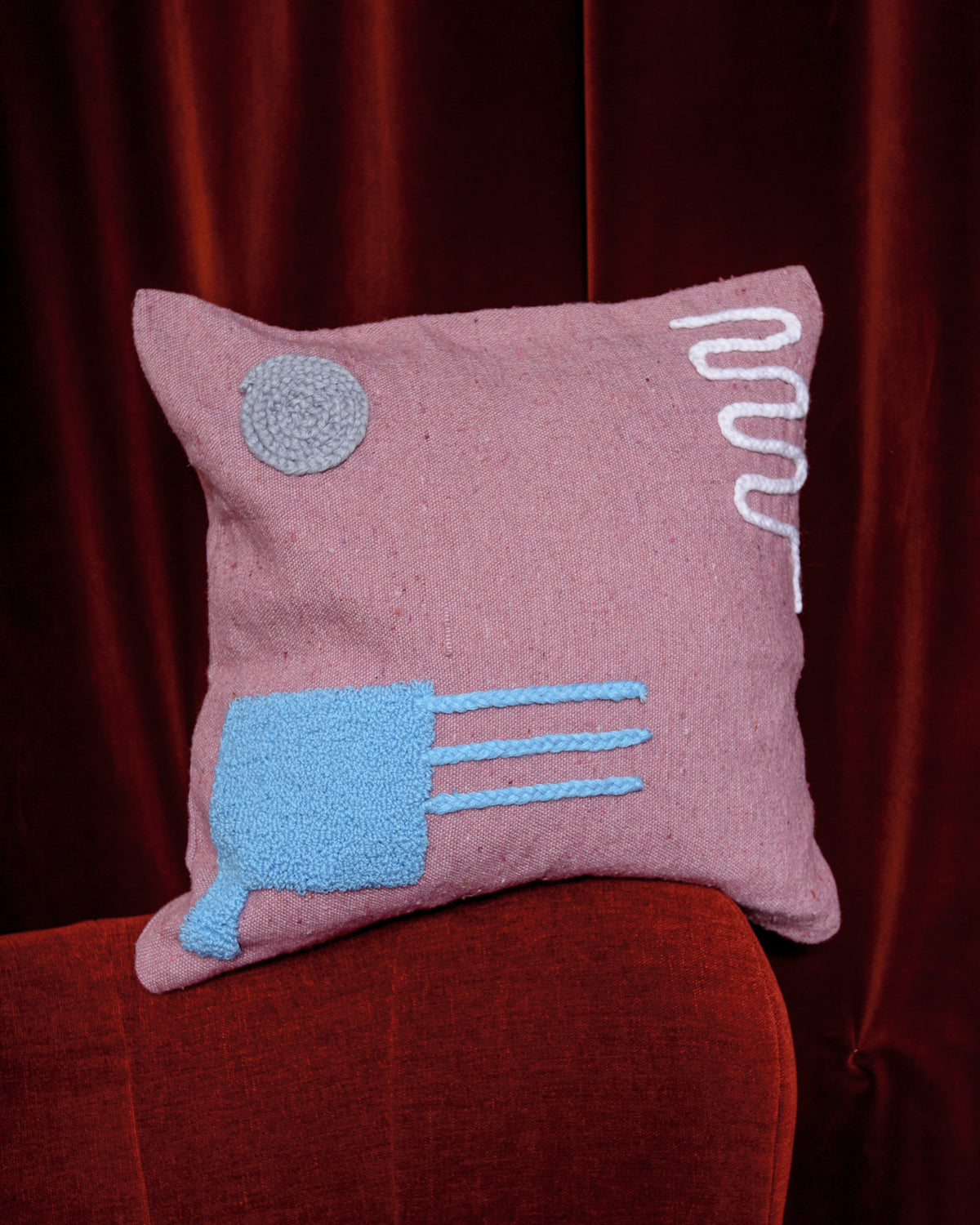 Pink wool cushion with embroidered design sitting on edge of red velvet armchair with red velvet curtain behind.