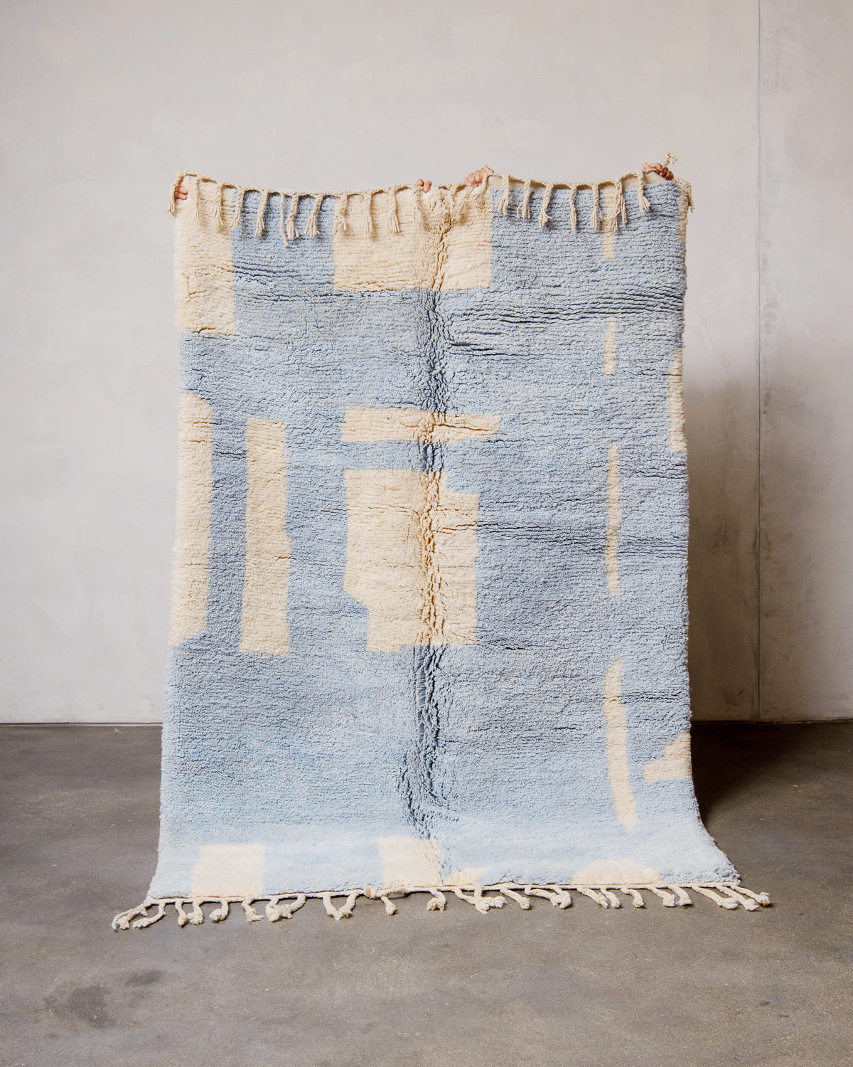 Berberlin Finest Rug made of cream and baby blue wool being held up in front of a white wall and concrete floor. 