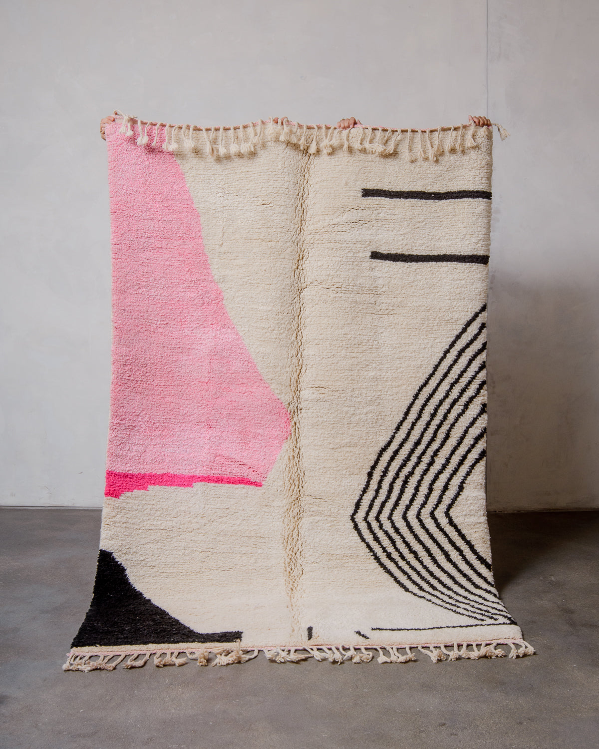 Berberlin Finest Rug made of cream wool with a pink and black design being held up in front of a white wall and concrete floor. 