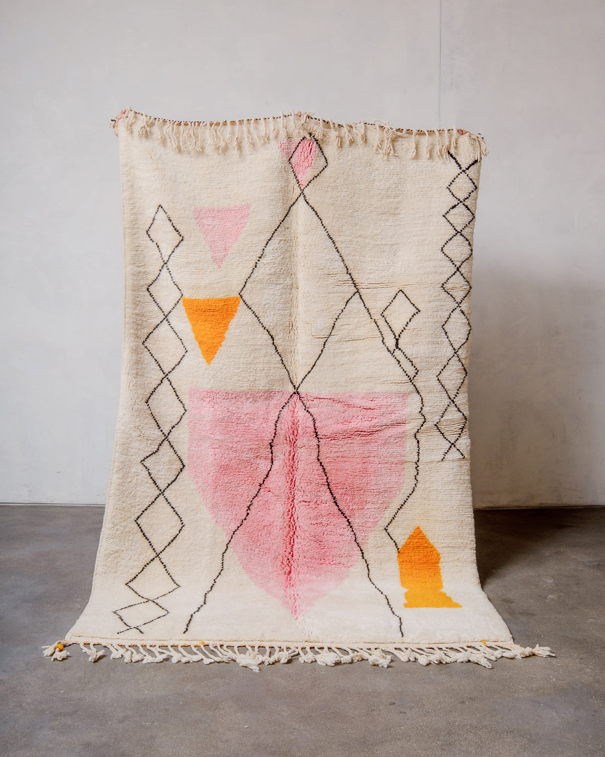 Berberlin Finest Rug made of plush cream wool with a design in pink, orange and black being held up in front of a white wall and concrete floor. 