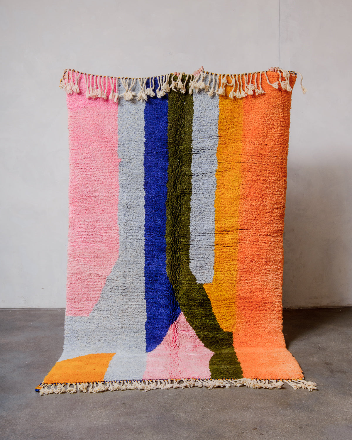Berberlin Finest Rug made of heavy wool pile in bright colours being held up in front of a white wall and concrete floor. 