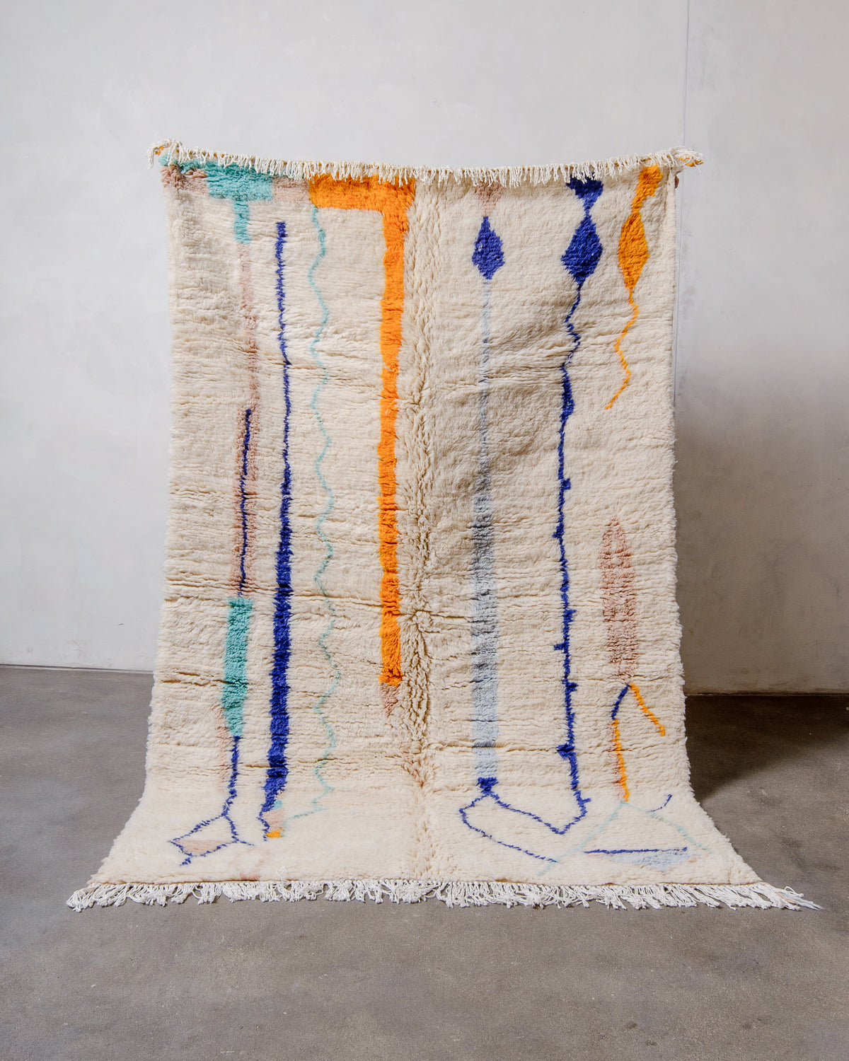 Berberlin Finest Rug made of plush wool in cream, orange, turquoise, blue and old rose, being held up in front of a white wall.