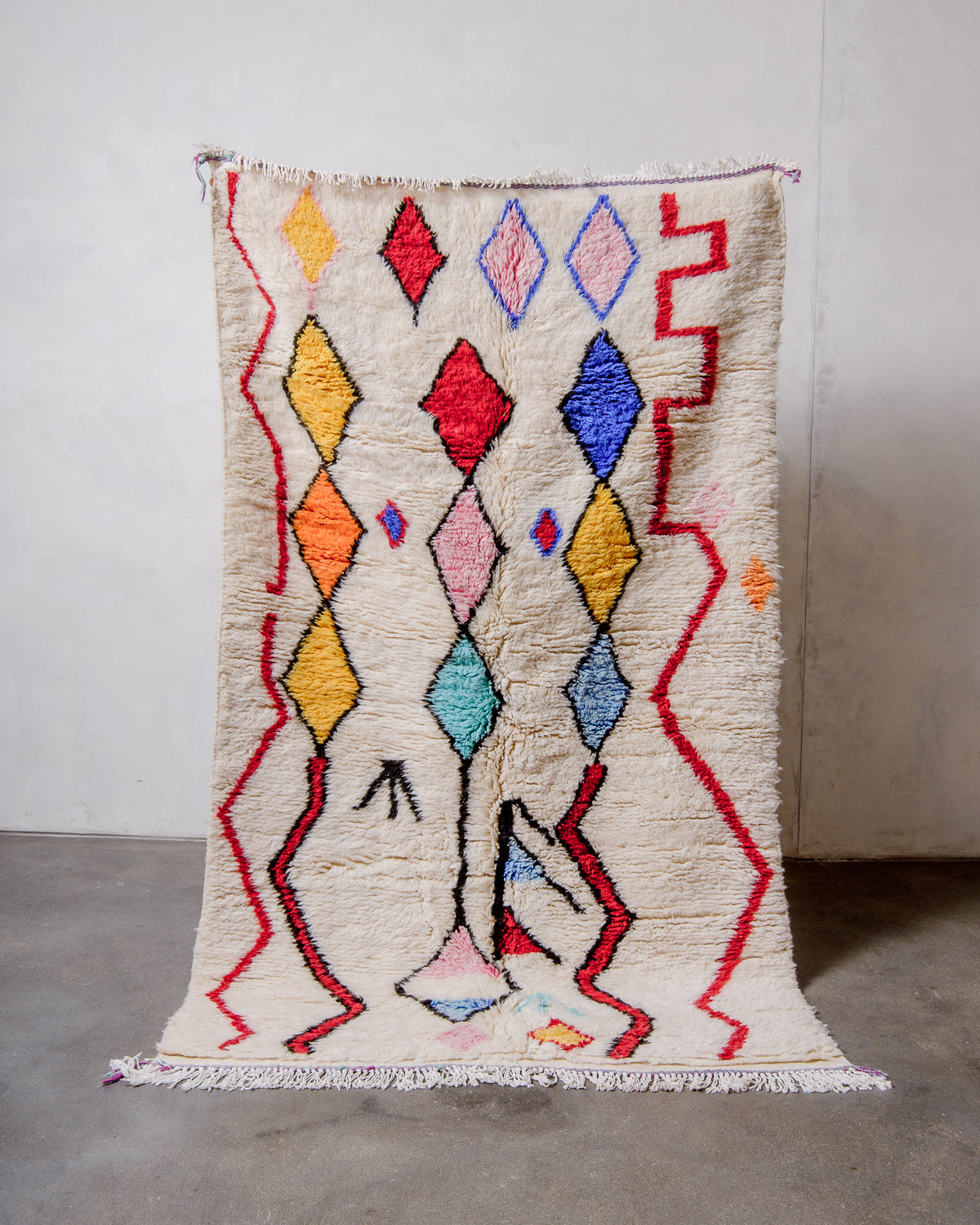 Berberlin Finest Rug made of plush cream wool with a colourful design being held up in front of a white wall and concrete floor. 