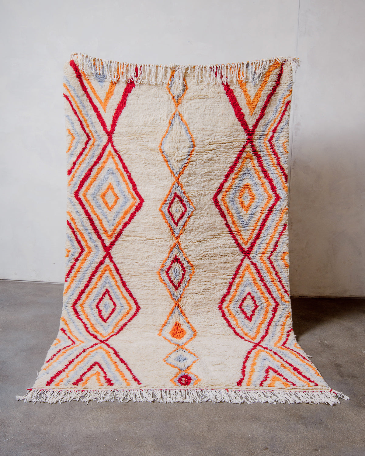 Berberlin Finest Rug made of plush cream wool with a colourful design in red, orange and light blue, being held up in front of a white wall.