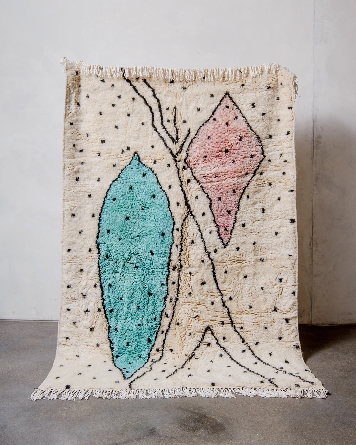 Berberlin Finest Rug made of plush cream wool with a cool design in including large pink and turquoise blue forms, and black polka dots, being held up in front of a white wall and concrete floor. 