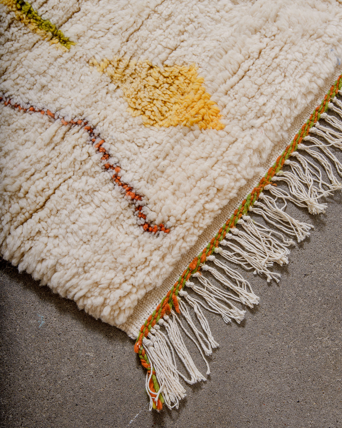 Corner of Berber rug made of thick, fluffy wool pile and finished with green and orange woven ends and white cotton tassels. 