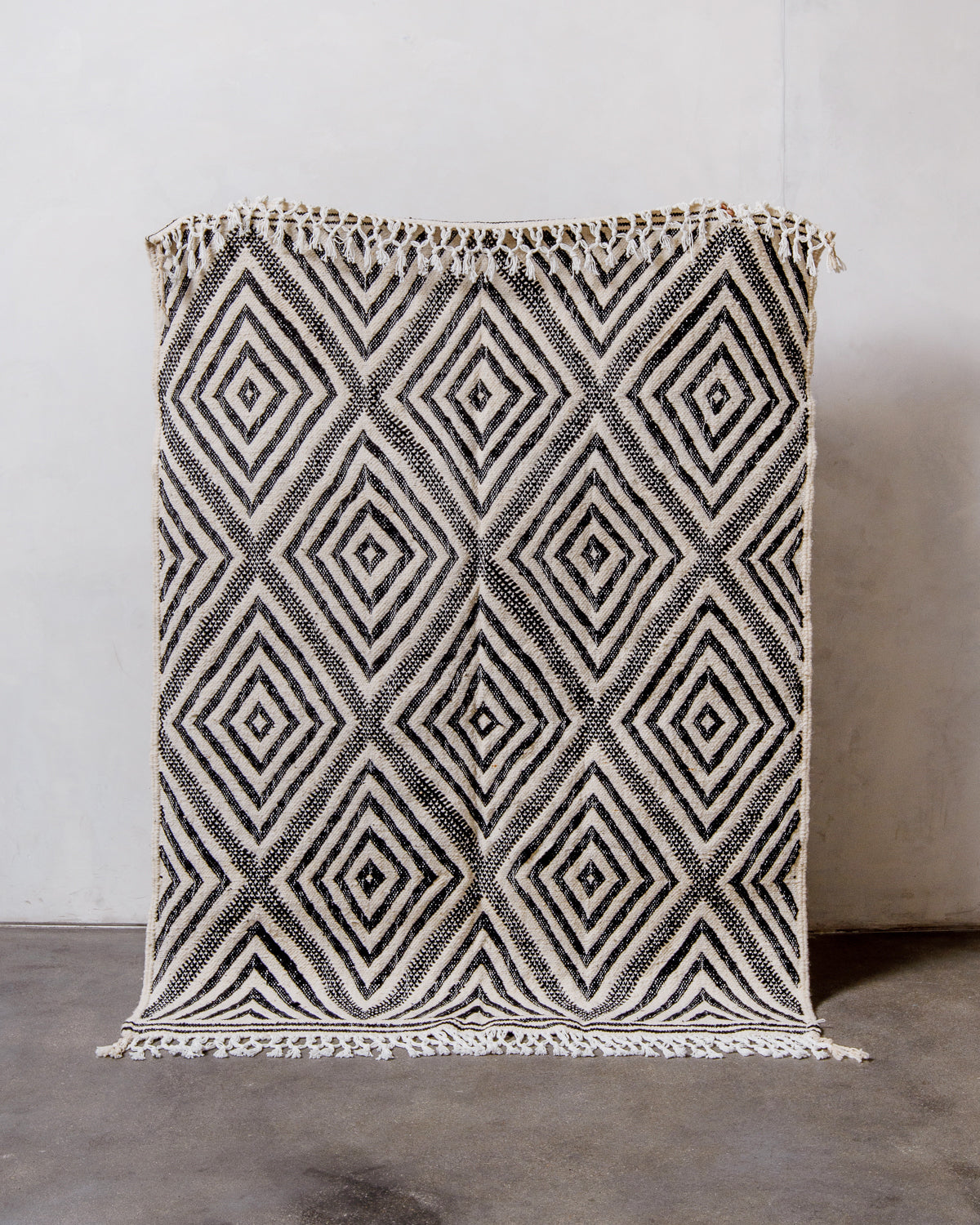 Berberlin Kelim rug with black and white diamond pattern, being held up in front of a white wall. 