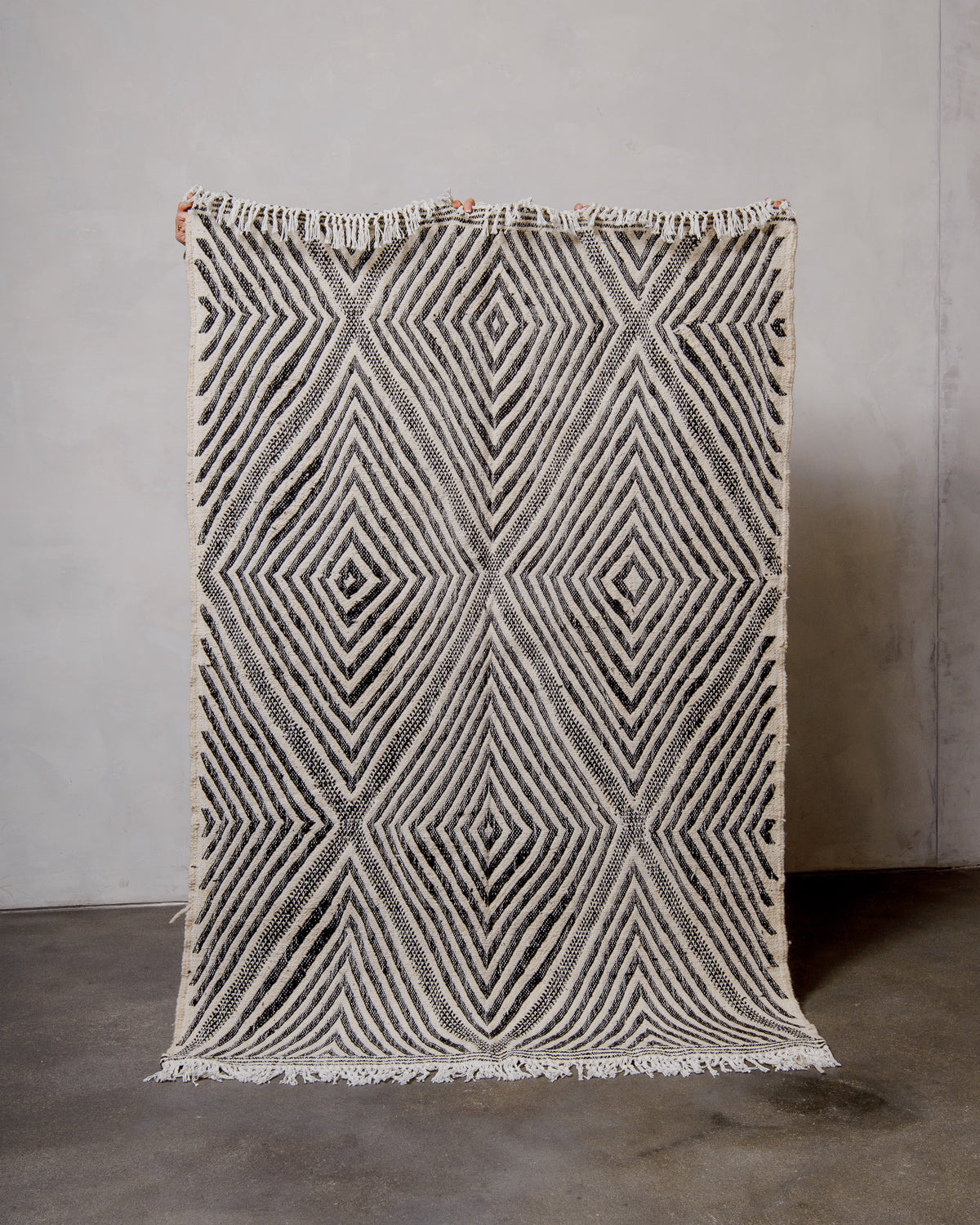 Berberlin Kelim rug made of black and white wool being held up in front of a white wall. 
