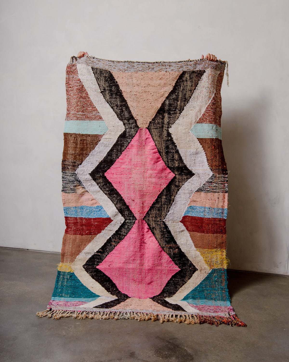 Berberlin vintage kelim rug made of recycled materials being held up by two hands in front of a white wall and concrete floor. 