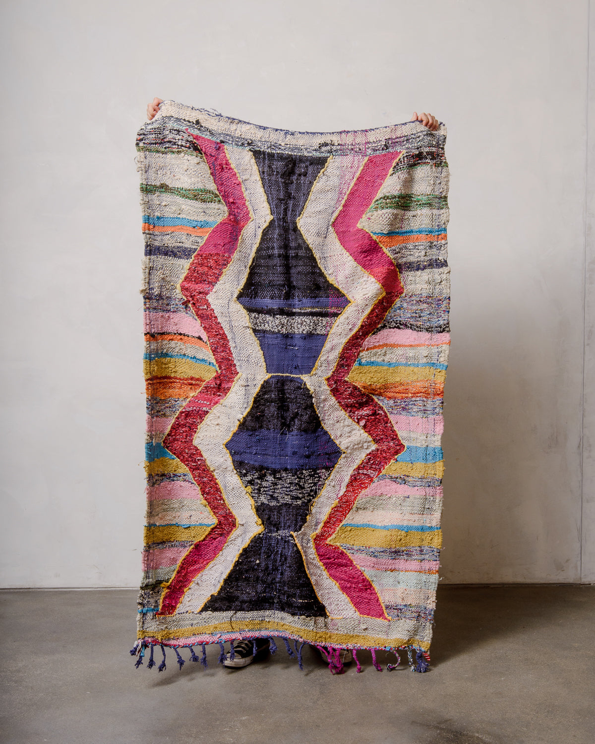 Berberlin vintage kelim rug made of recycled materials being held up by two hands in front of a white wall and concrete floor. 