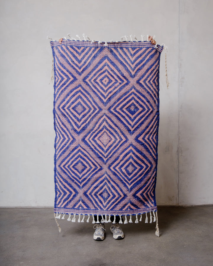 Small Berberlin Kelim Rug in blue and pink, being held up in front of a white wall.