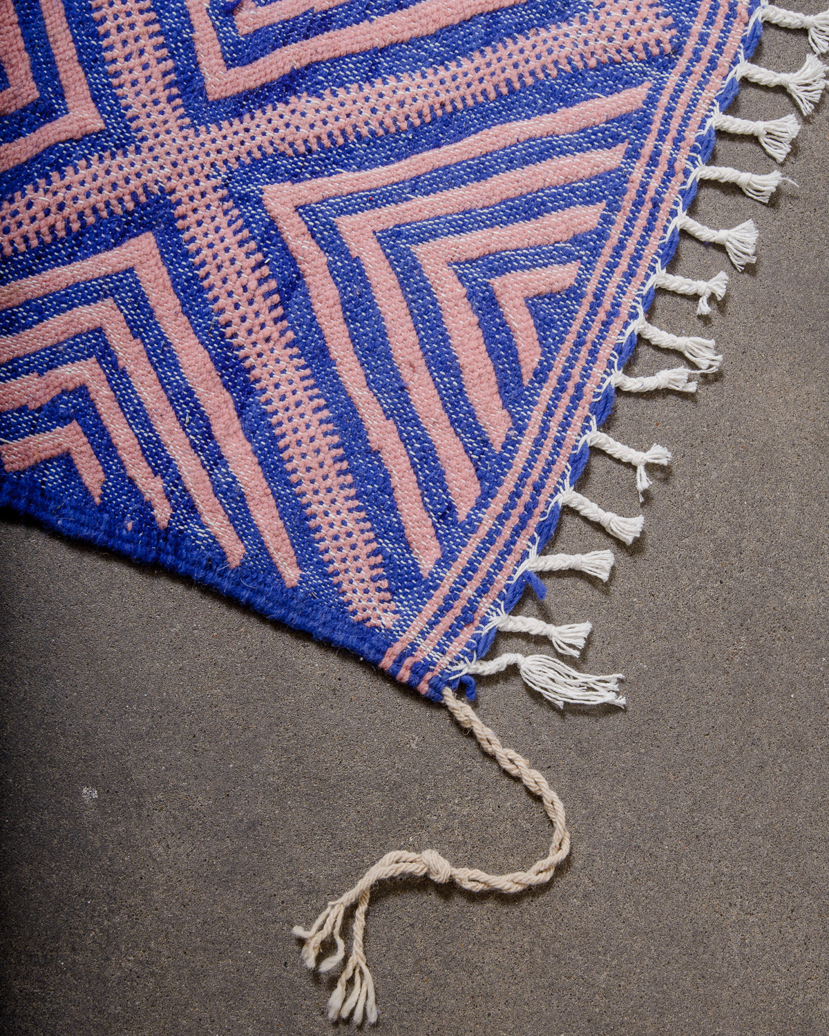 Corner of Kelim Rug in blue and pink with white cotton tassels, laying on concrete.