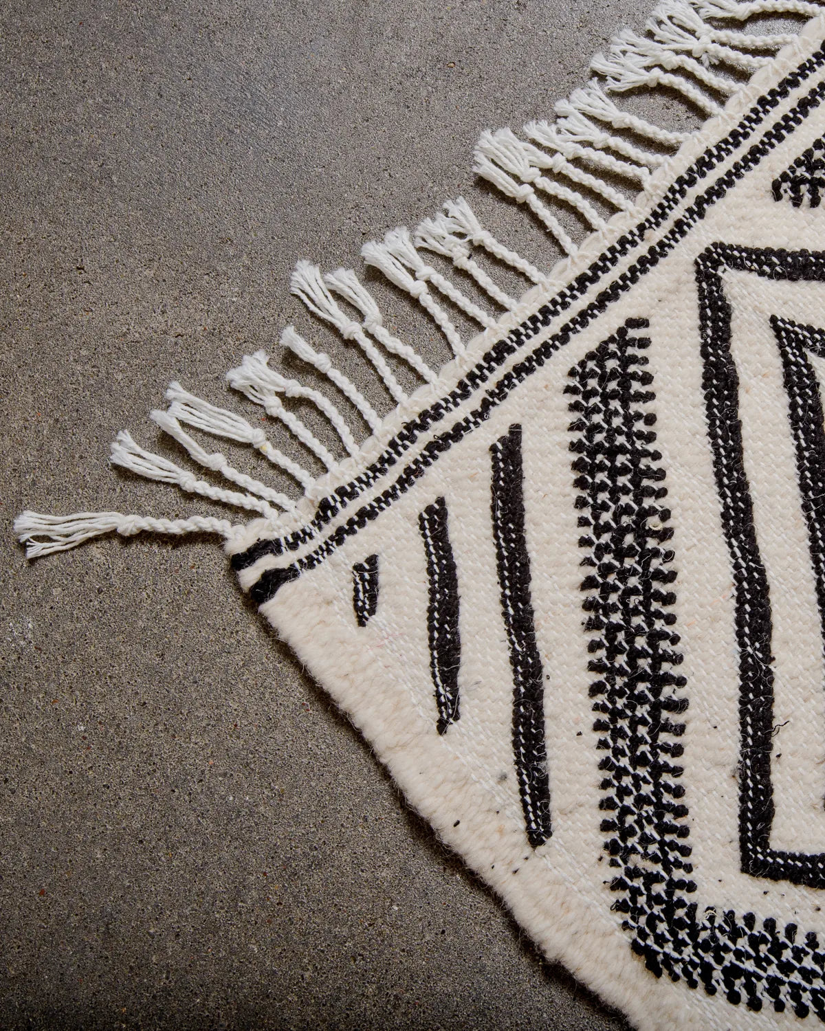 Corner of Kelim Rug in black and white with white cotton tassels, laying on concrete.