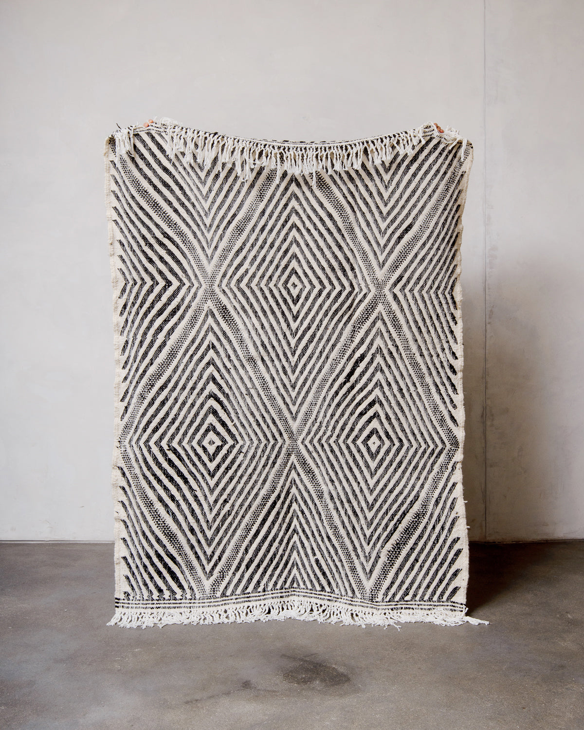 Berberlin Kelim rug made of black and white wool being held up in front of a white wall. 