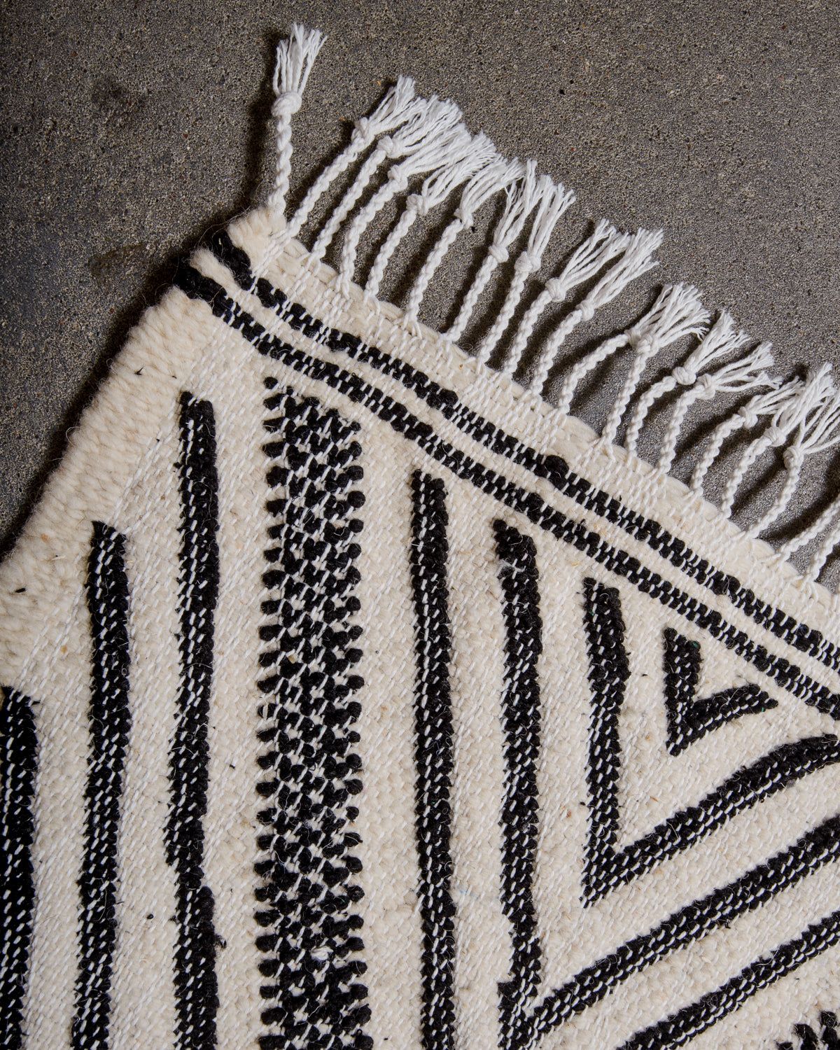 Corner of Kelim Rug in black and white with white cotton tassels, laying on concrete.
